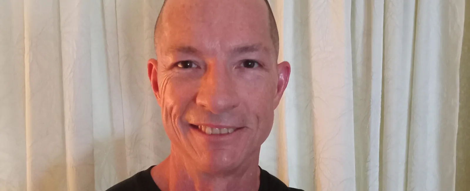 Smiling man with a mohawk haircut wearing a black YouFit Gyms t-shirt in front of white curtains.