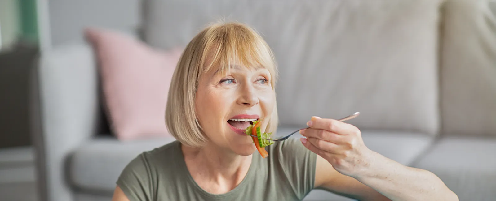Middle-aged woman eating a fresh salad while sitting next to a blue exercise ball at home.