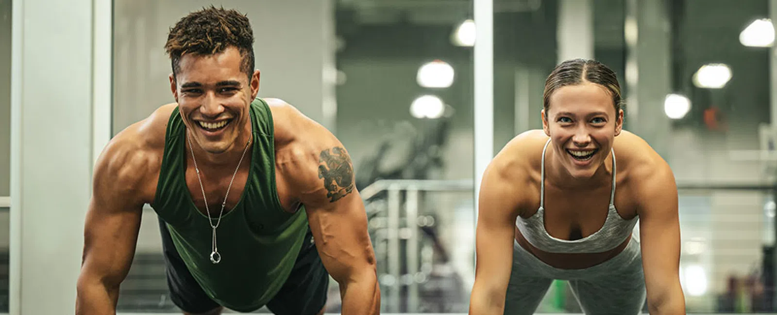 Smiling man and woman doing push-ups together in a gym with fitness equipment in background