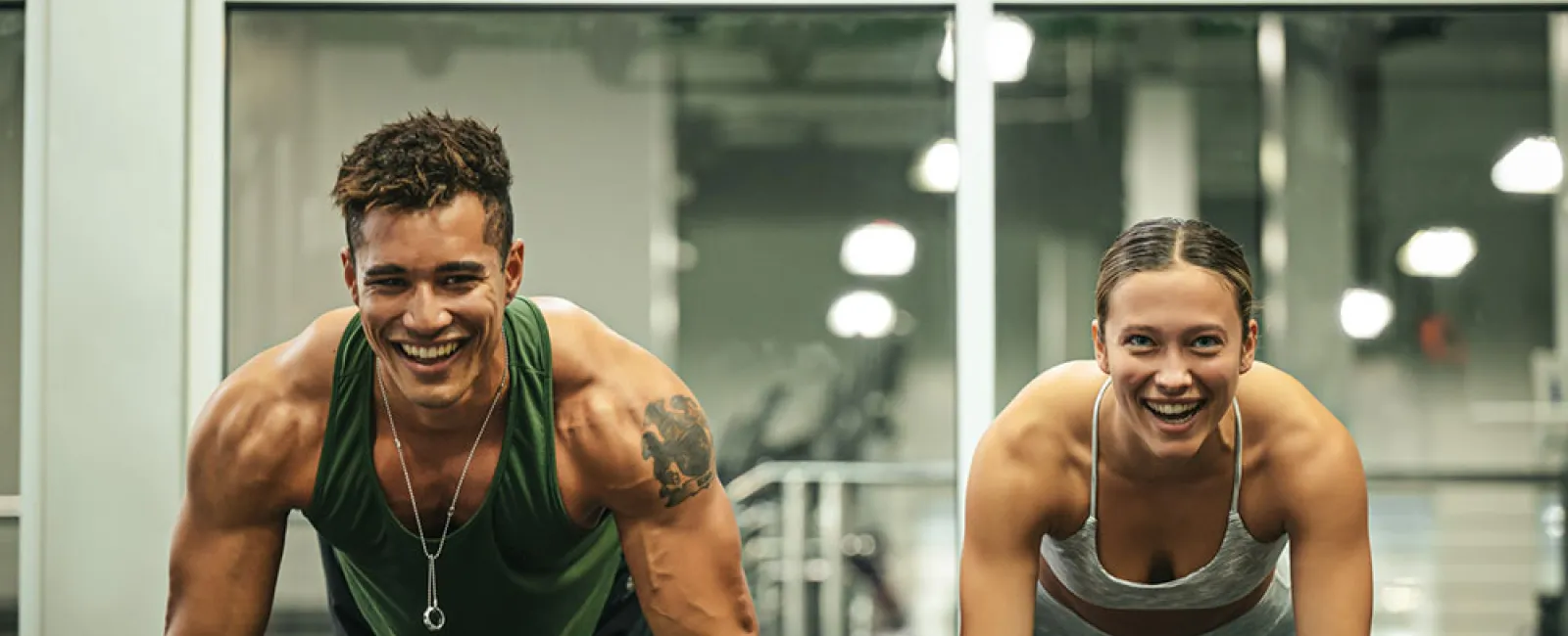 Young man and woman doing push-ups together in a gym with smiles, wearing athletic clothing.