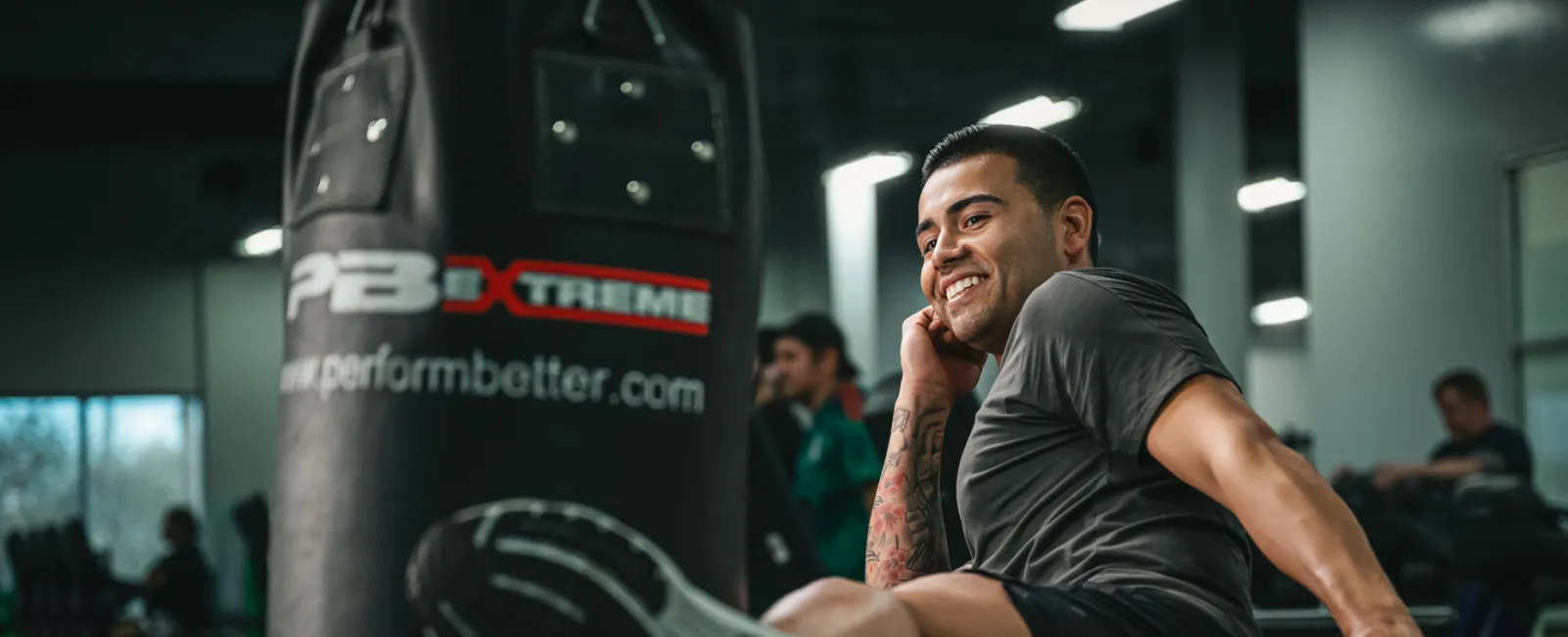 Man in athletic wear kicking a punching bag in a gym with a focused and smiling expression.