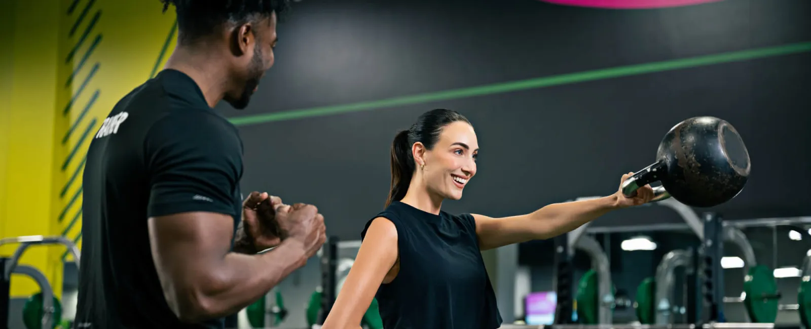 Female lifting kettlebell with male trainer coaching her in gym with weight racks and colorful plates in background