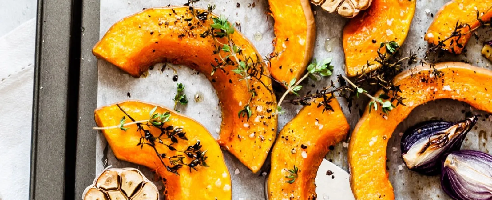 Roasted pumpkin slices with garlic, red onion, and fresh thyme on a baking tray seasoned with pepper and salt.
