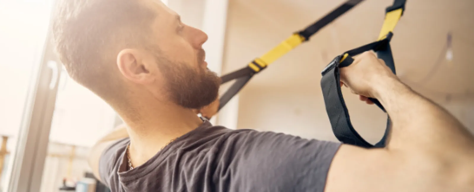 Man with beard exercising using suspension trainer straps in a bright indoor gym space.
