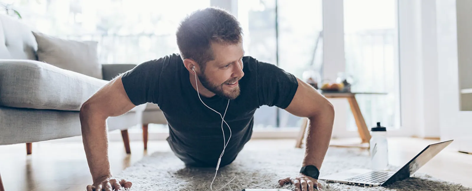 Man doing push-ups on a rug while watching a laptop and listening to music with earphones at home.