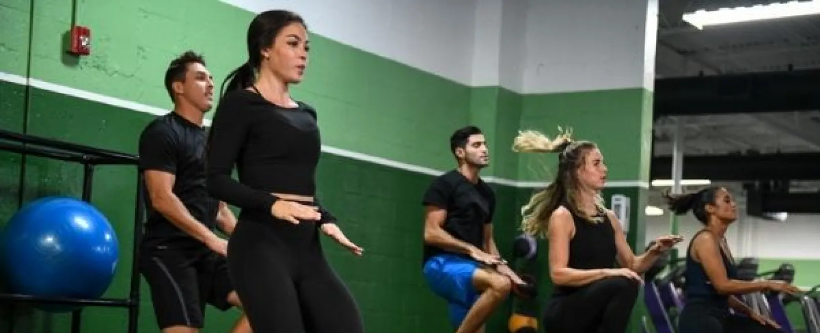 Group of five people exercising with high knees in a gym with green and white walls and fitness equipment.