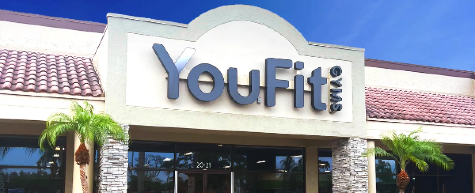 YouFit Gyms storefront with stone pillars, palm trees, and clear blue sky in a shopping plaza.