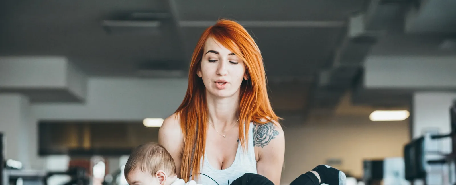 Young woman with red hair exercising by lifting a baby in a gym setting with workout equipment in the background