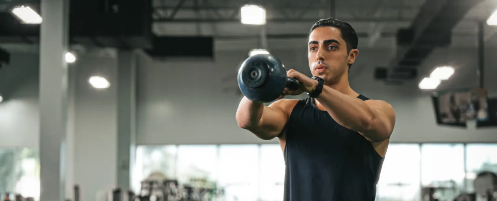 Man performing kettlebell exercise in modern gym wearing black tank top and shorts