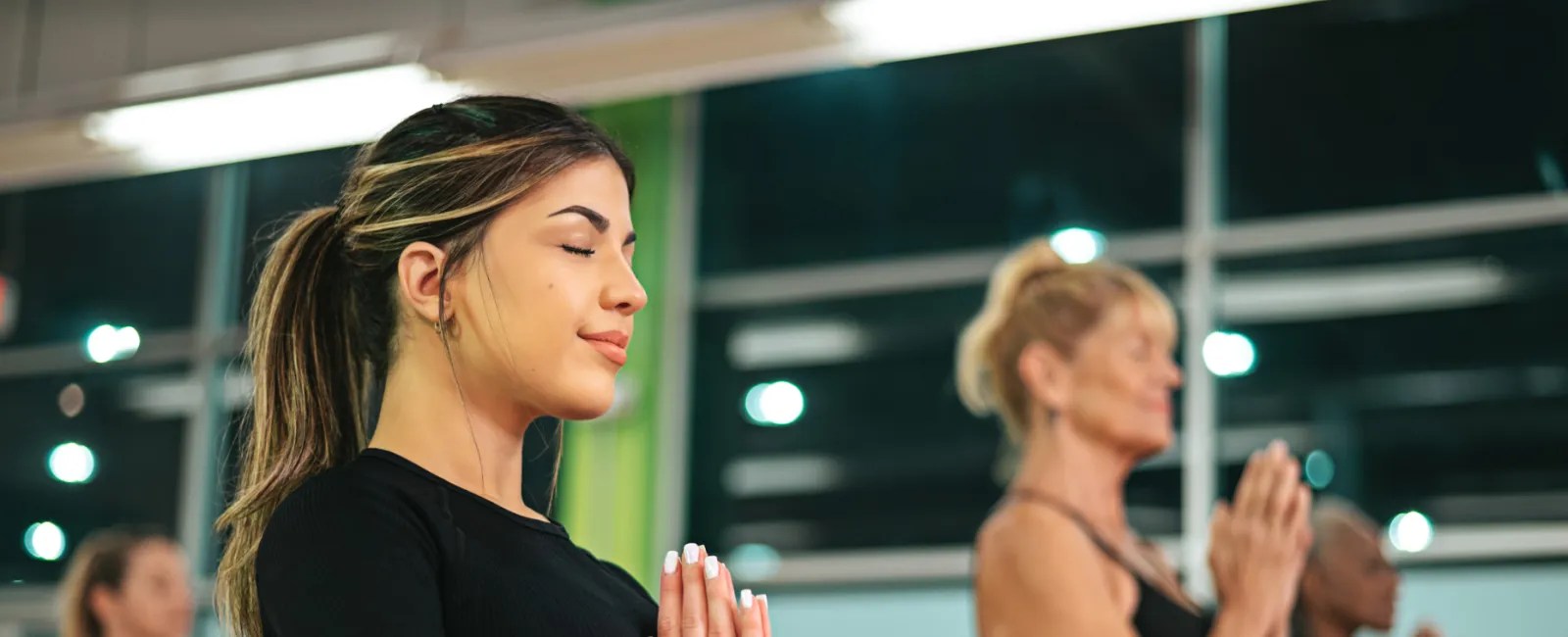 Women practicing yoga in a studio with hands in prayer pose and eyes closed, focusing on mindfulness and relaxation.