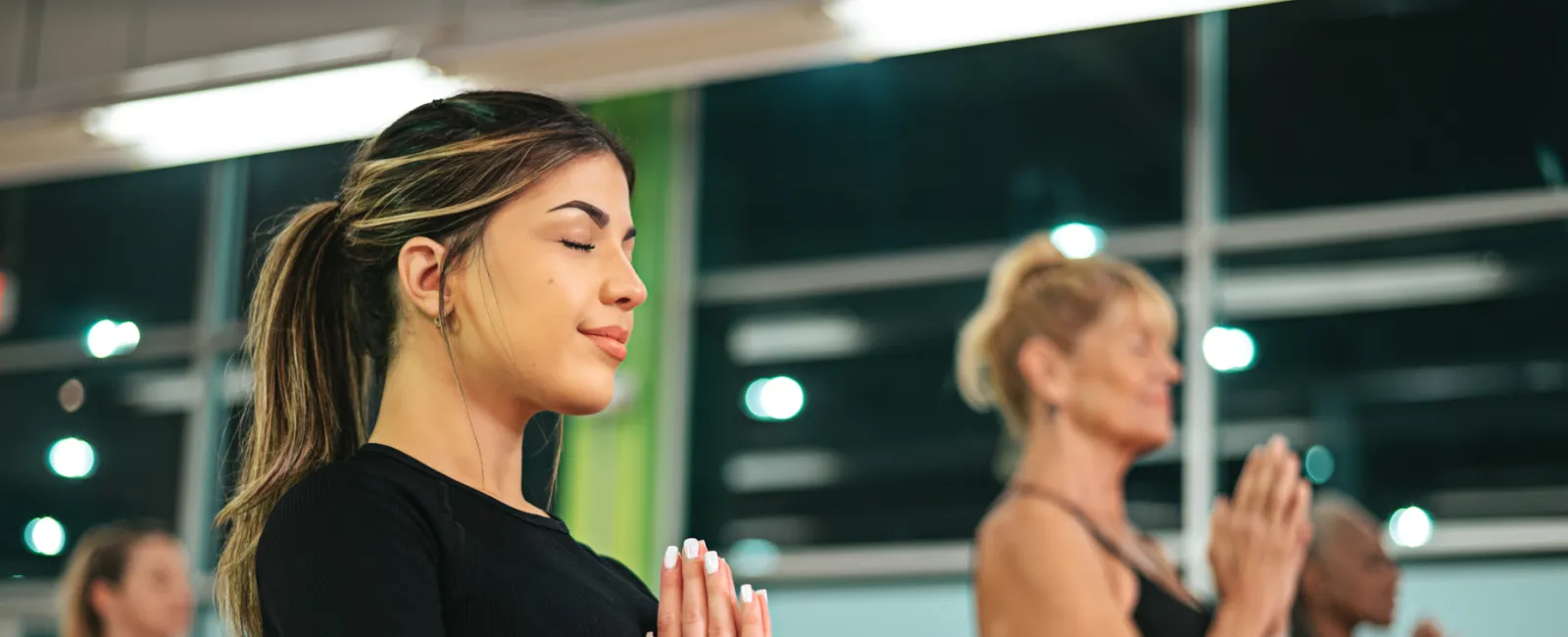 Women practicing yoga in a studio with hands in prayer pose and eyes closed, focusing on mindfulness and relaxation.