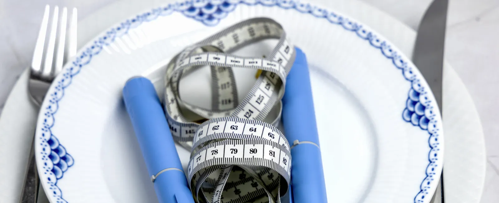 Blue insulin pens and a white measuring tape on a white plate with blue patterns, fork and knife beside.