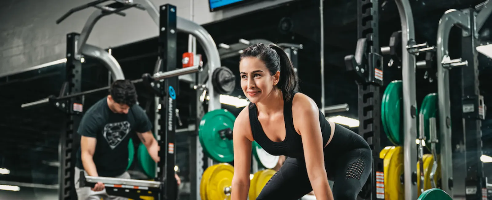 Woman lifting barbell with weights in gym while a man adjusts equipment in the background