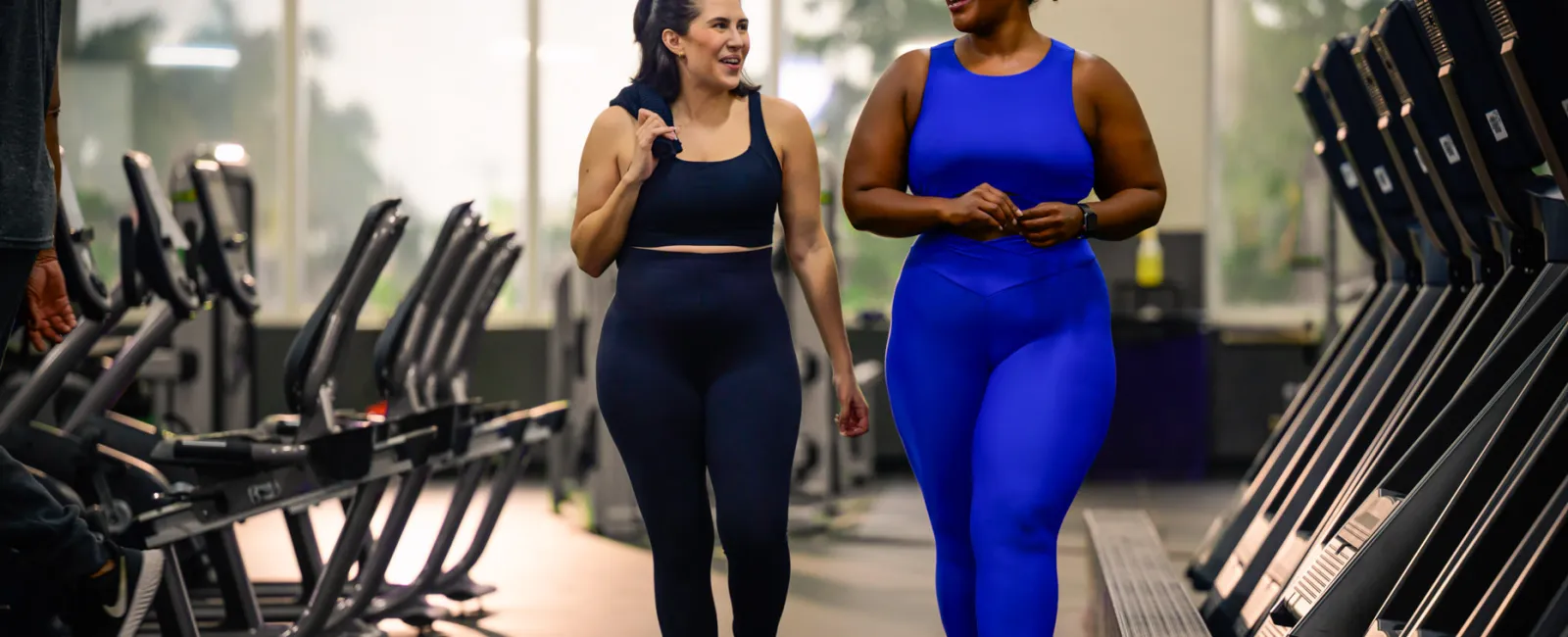Two women in activewear walking and smiling in a gym surrounded by cardio equipment and natural light.