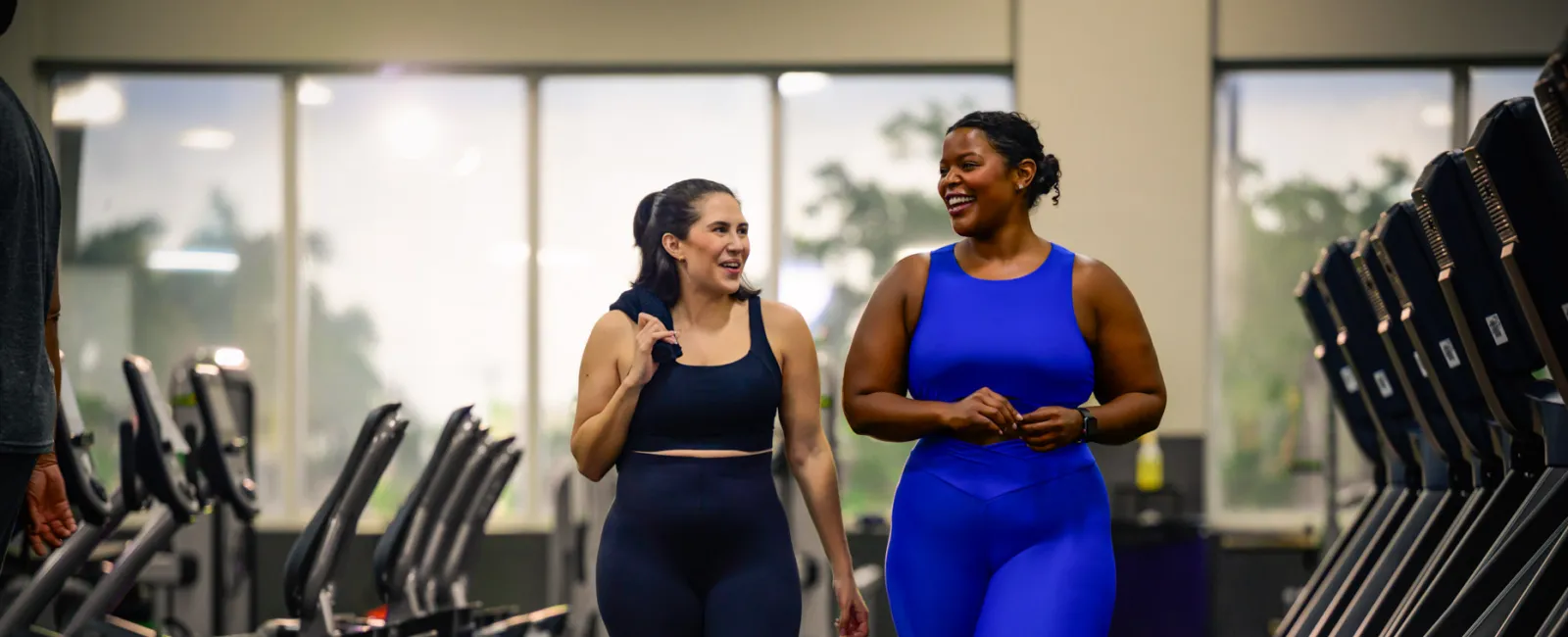 Two women in activewear walking and smiling in a gym surrounded by cardio equipment and natural light.