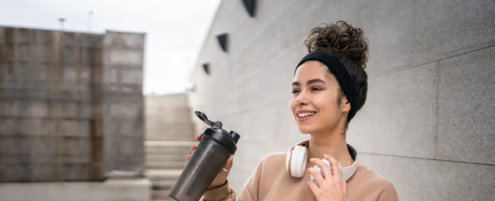 Smiling woman in casual wear holding a water bottle and white headphones outdoors by concrete walls.