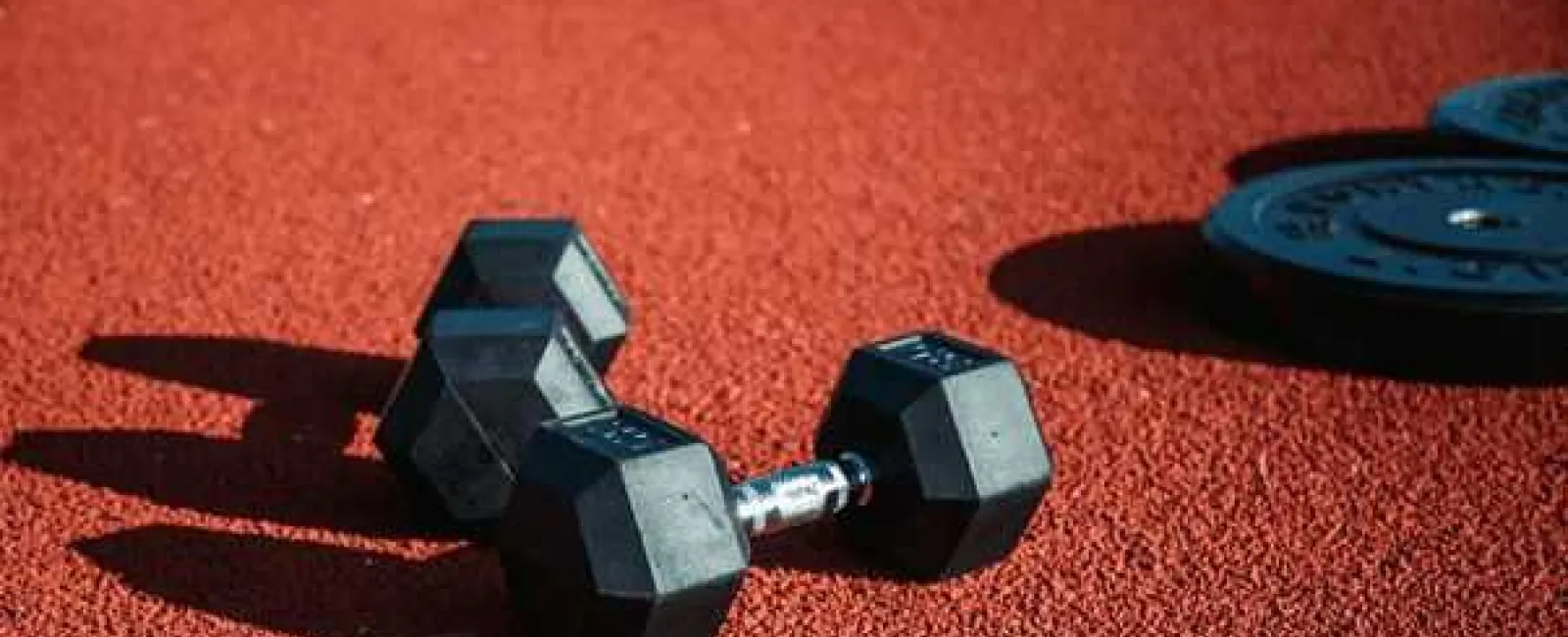 Black hex dumbbells and weight plates on a red textured gym floor with jump rope.