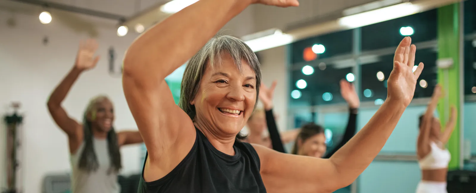 Smiling mature woman in black top leading group exercise class indoors with participants raising arms and moving.