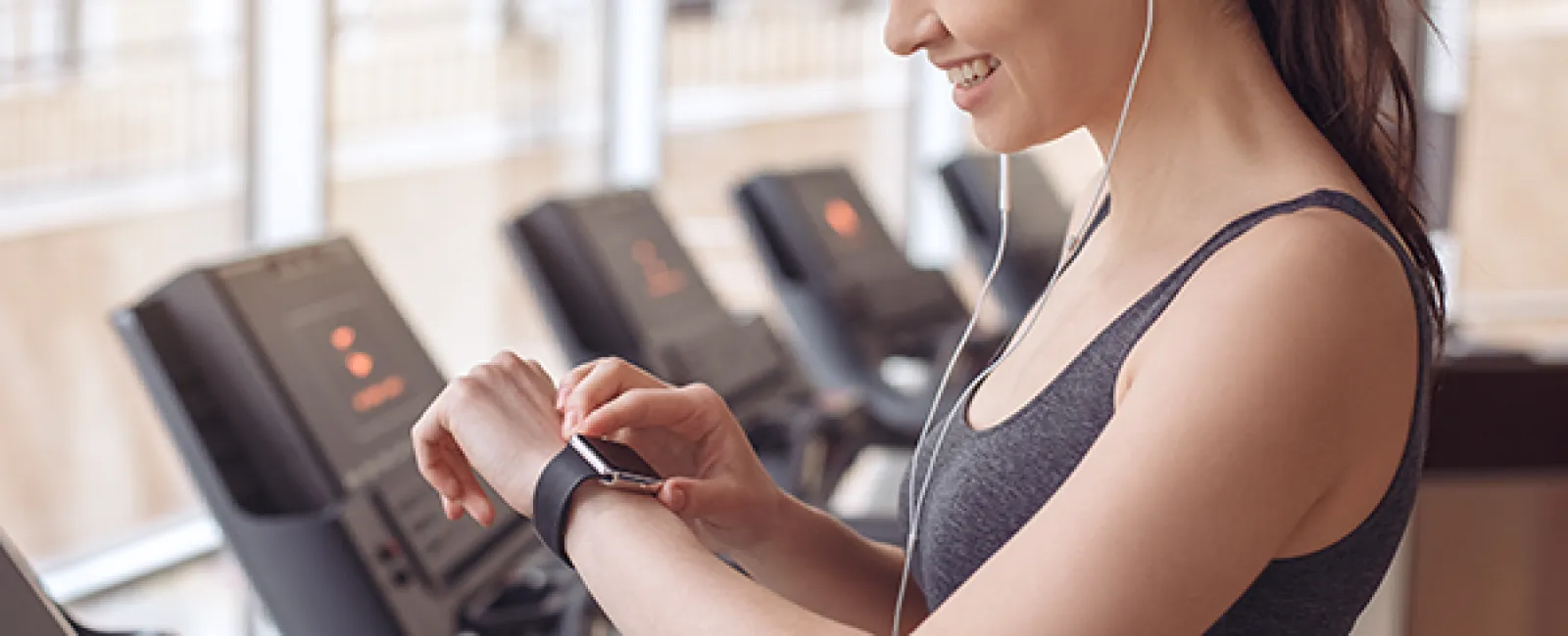 Smiling woman in gym adjusting smartwatch while wearing earphones near treadmills with large windows.