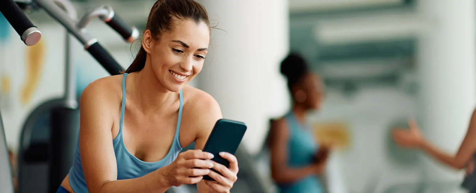 Smiling woman in blue workout clothes using smartphone while sitting on gym bench with equipment in background