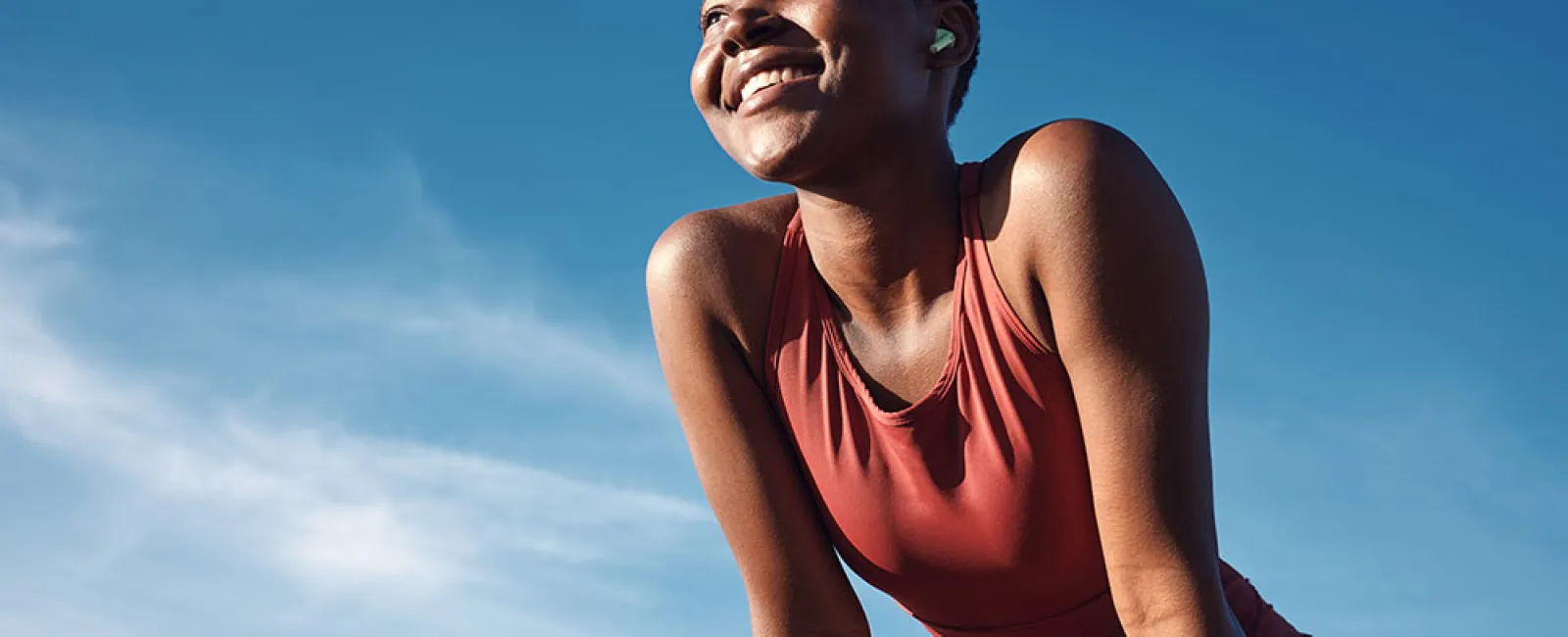 Smiling woman in red workout clothes wearing wireless earbuds outdoors under clear blue sky.