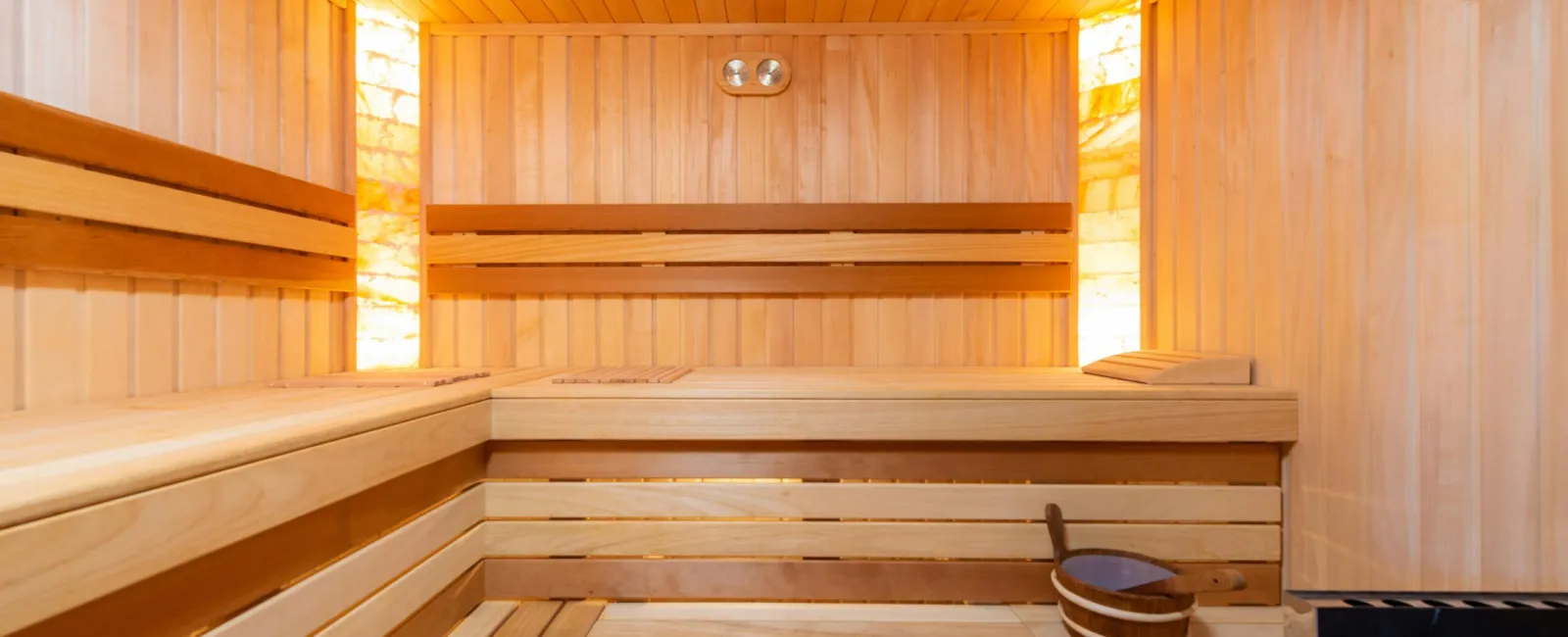 Interior of a wooden sauna with benches, backrests, warm lighting, and a sauna bucket with ladle.