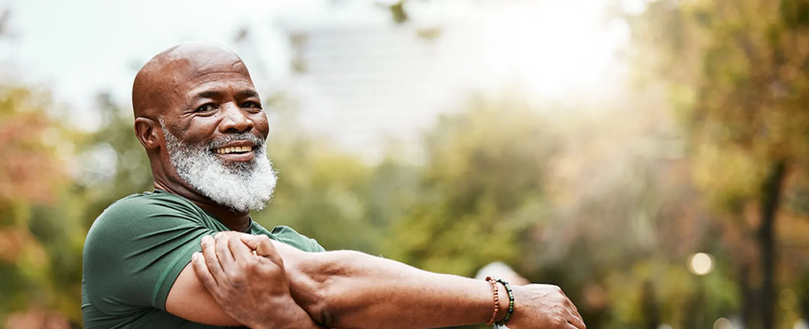 Smiling elderly man stretching his arm in a park with people exercising in the background on a sunny day