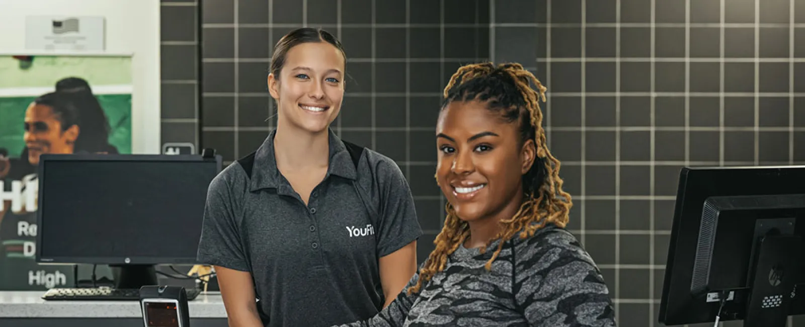 Smiling woman in workout clothes at a gym check-in counter with a friendly staff member behind the desk.