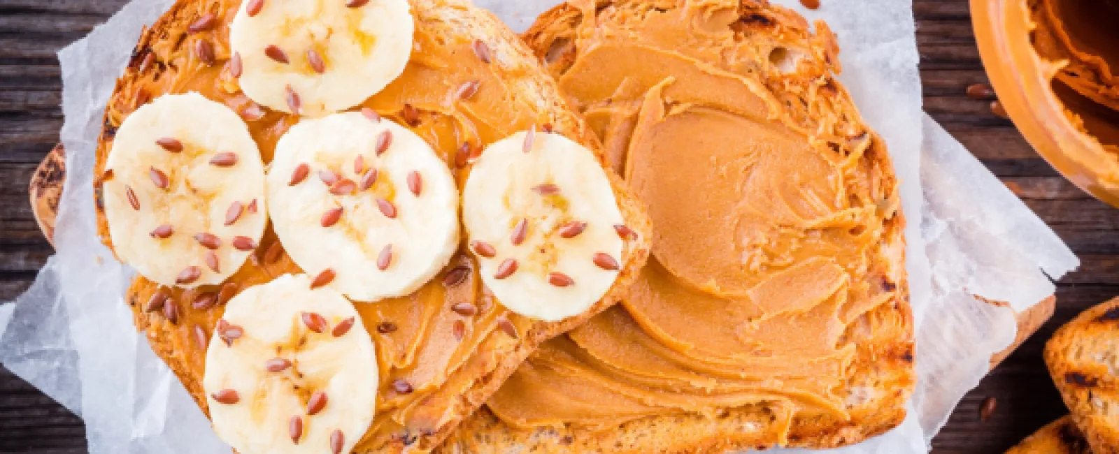 Peanut butter spread on toasted bread slices topped with banana slices and flax seeds on parchment paper.