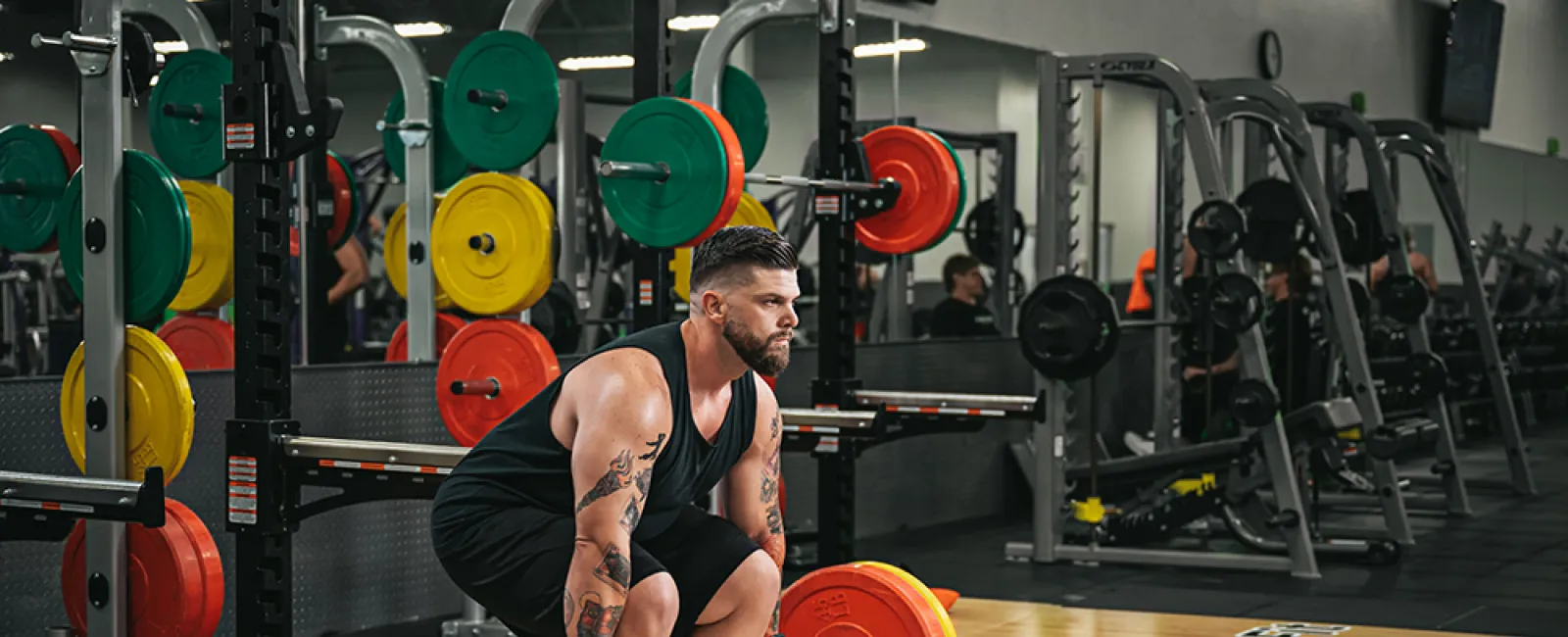 Muscular man with tattoos preparing to deadlift a barbell with colorful weight plates in a gym.