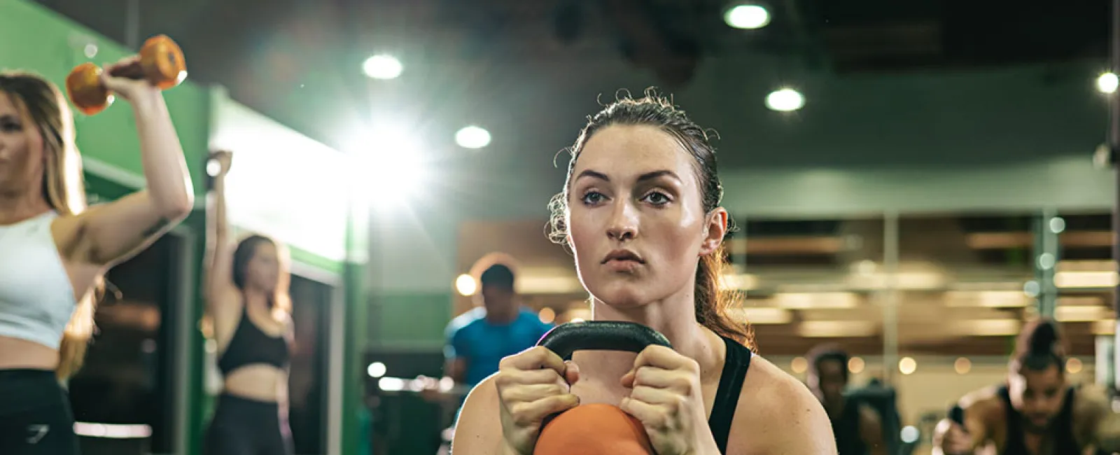 Focused woman holding a kettlebell in a gym surrounded by others exercising with weights and equipment