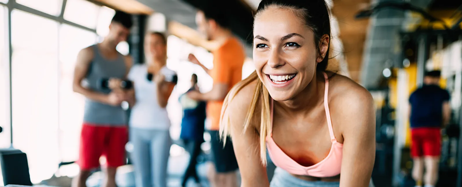 Smiling woman in pink sports bra leaning forward in gym with people training in the background