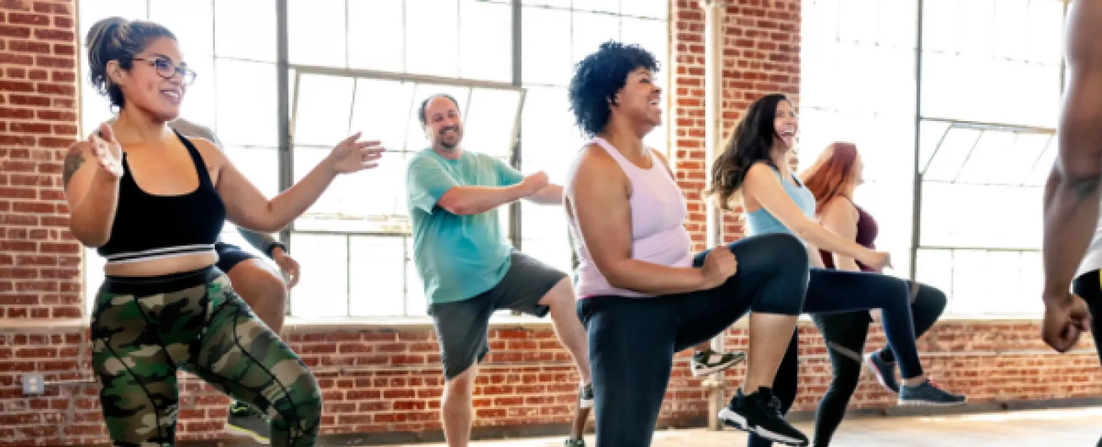 Group of diverse adults exercising and stretching together in a bright fitness studio with large windows.