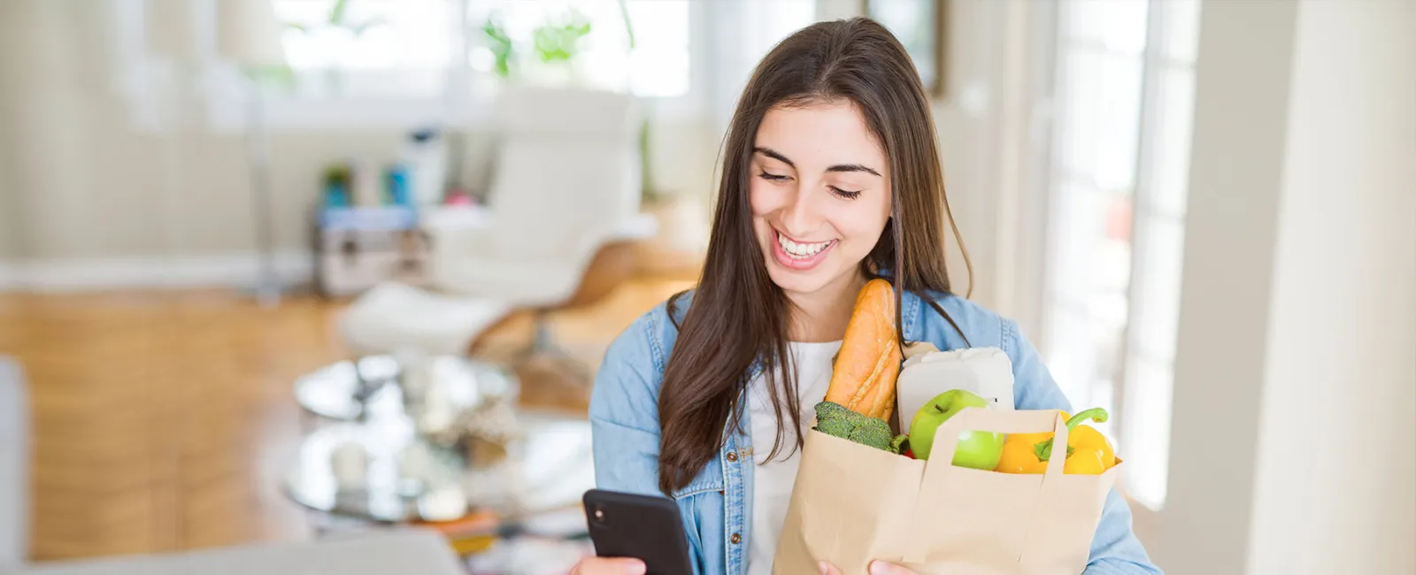 Smiling young woman holding groceries and checking phone in bright modern kitchen at home