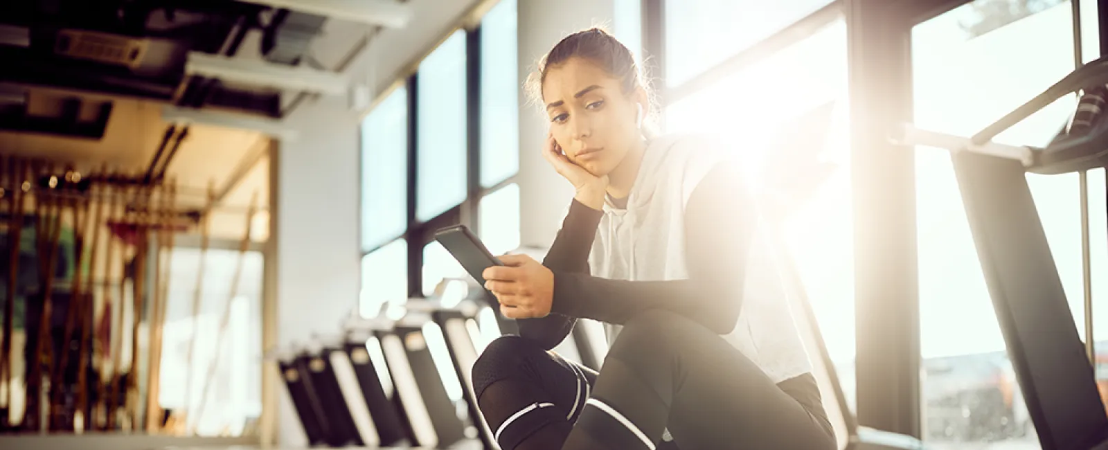 Young woman in athletic wear sitting on treadmill in gym looking at smartphone with thoughtful expression