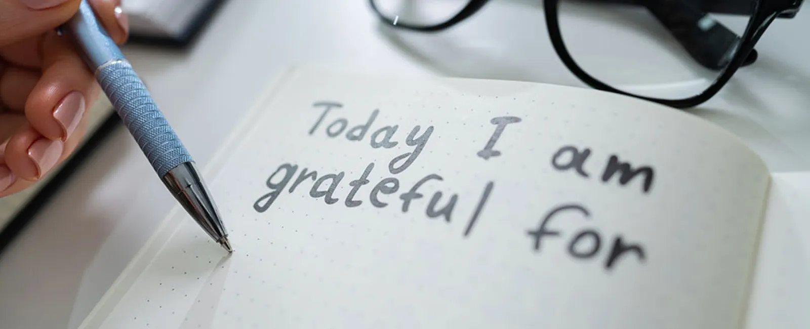 Hand writing gratitude journal entry saying Today I am grateful for with glasses and planner in the background