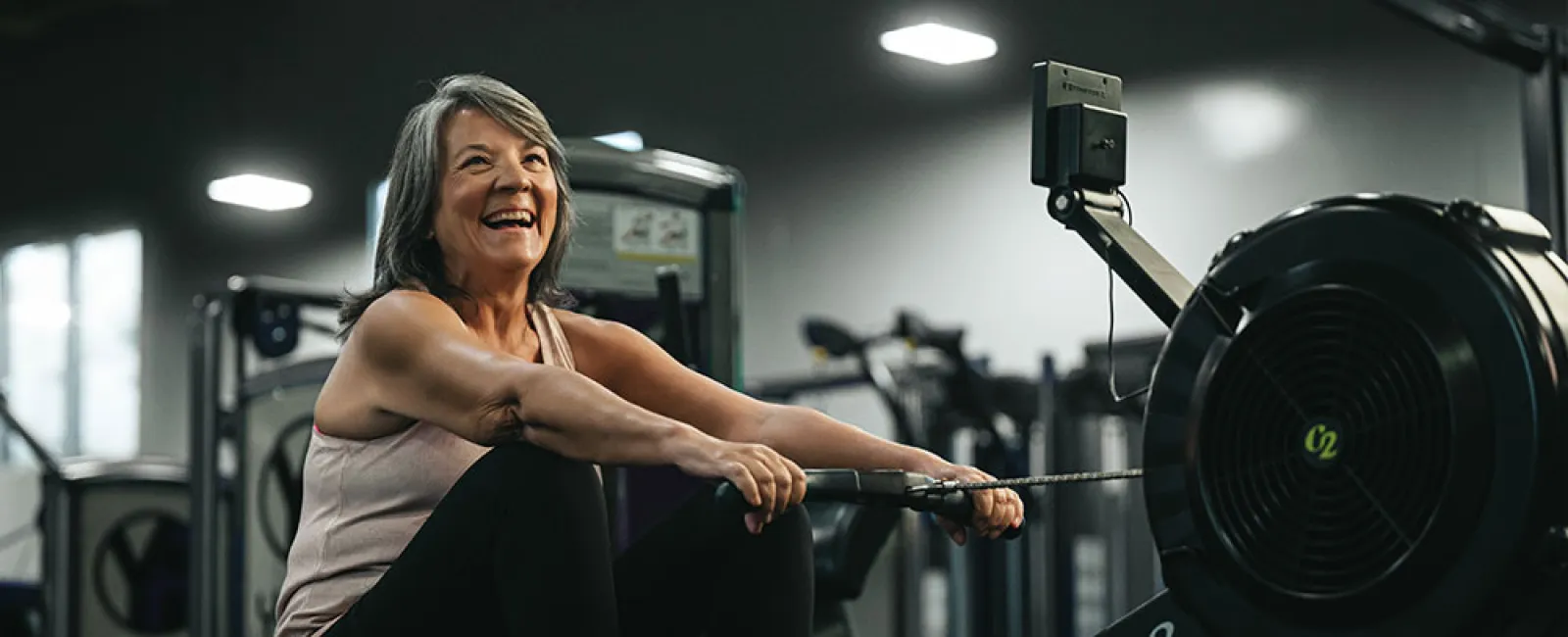 Mature woman smiling while using a rowing machine for exercise in a modern gym setting.