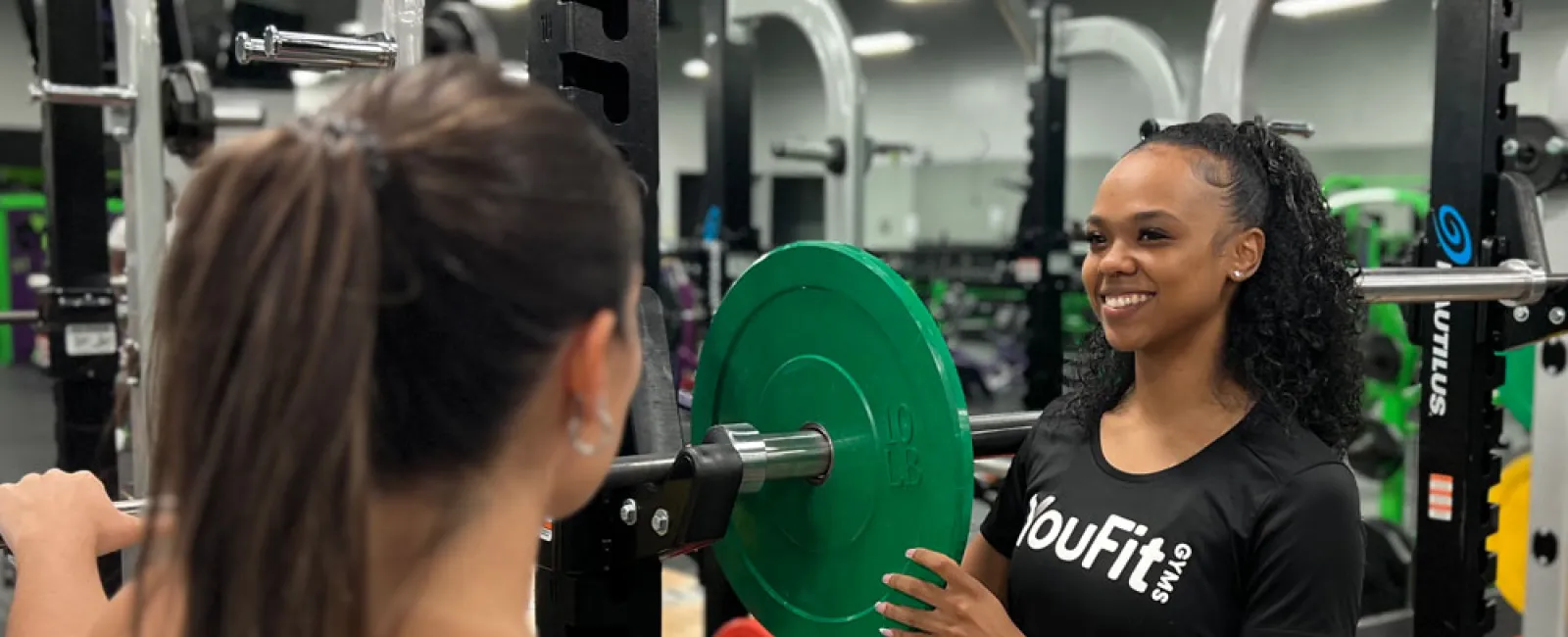 Personal trainer assisting woman with weightlifting at gym with green weight plate and fitness equipment.