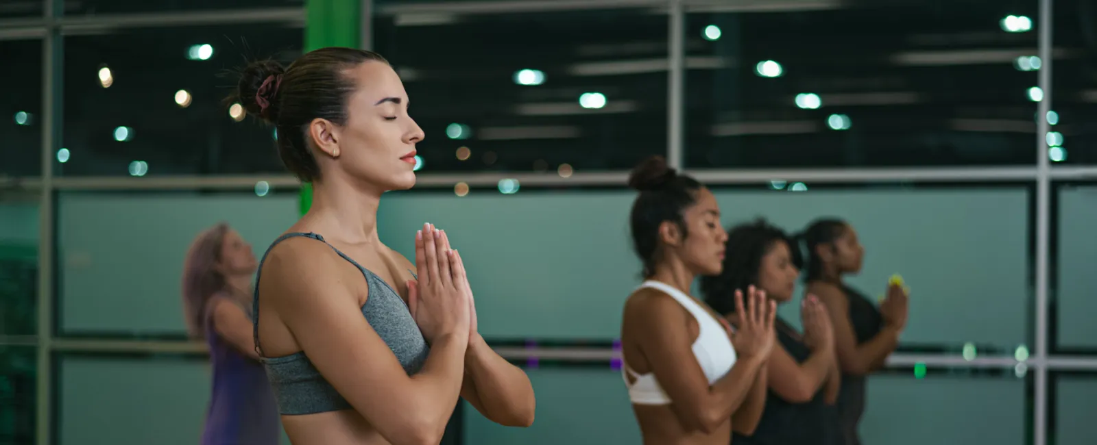 Group of women practicing yoga with hands in prayer pose inside a fitness studio.