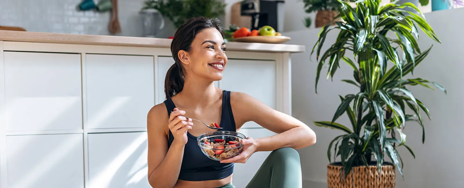 Woman in workout clothes sitting on floor eating a bowl of fruit, with dumbbells and a smoothie nearby in bright kitchen.