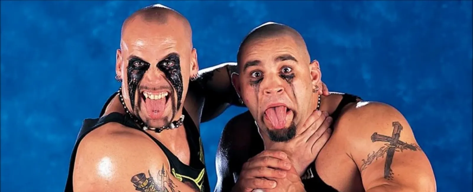 Two muscular men with face paint and tattoos pose playfully, one choking the other who sticks out his tongue.