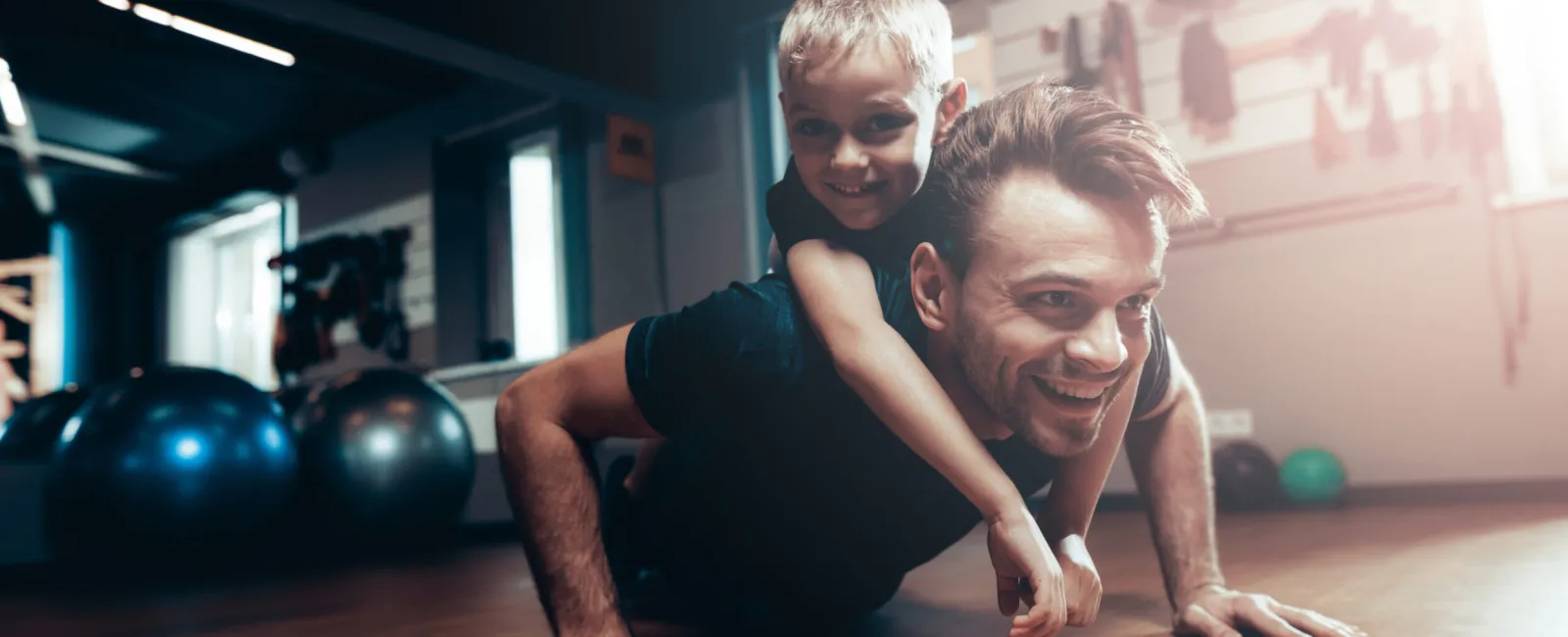 Man doing push-ups with a young boy on his back in a bright gym setting, both smiling and having fun.