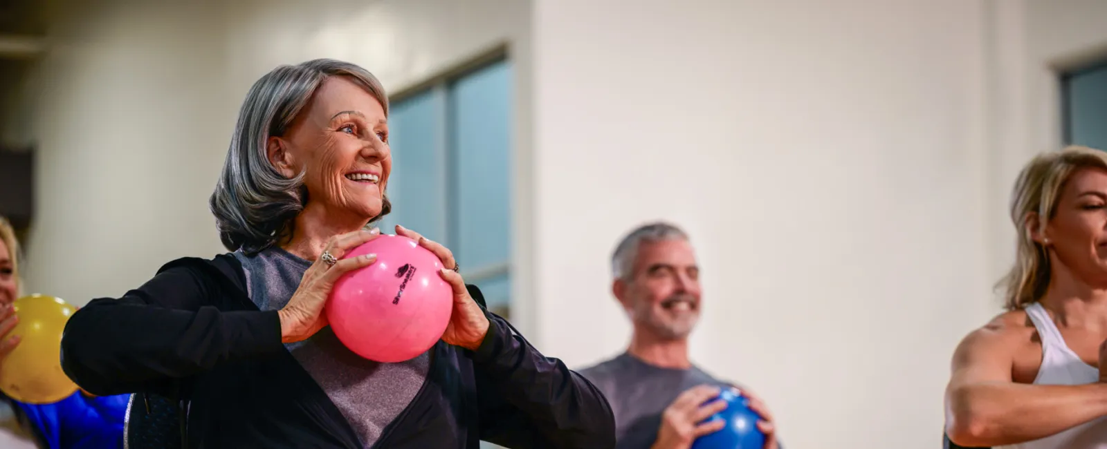 Seniors participating in a seated fitness class holding colorful exercise balls and stretching their legs.