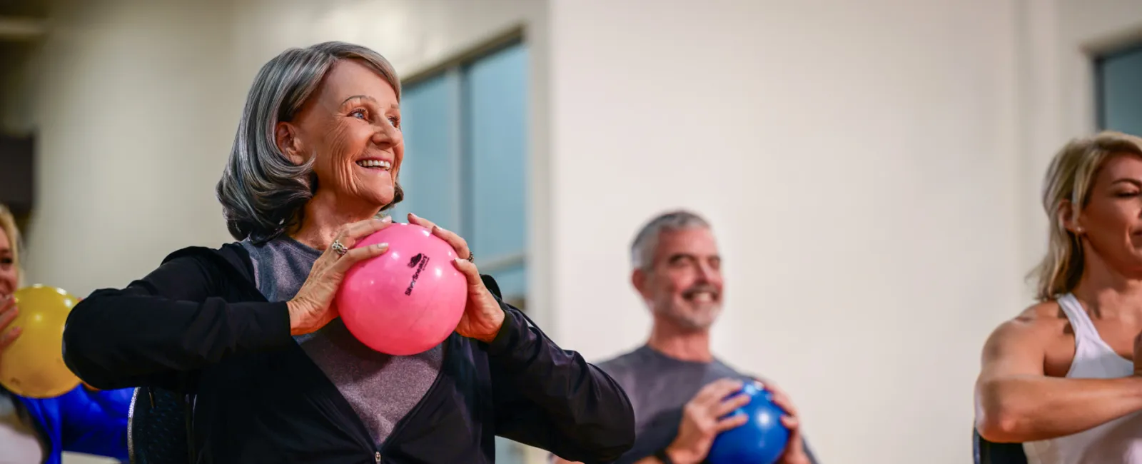 Seniors participating in a seated fitness class holding colorful exercise balls and stretching their legs.