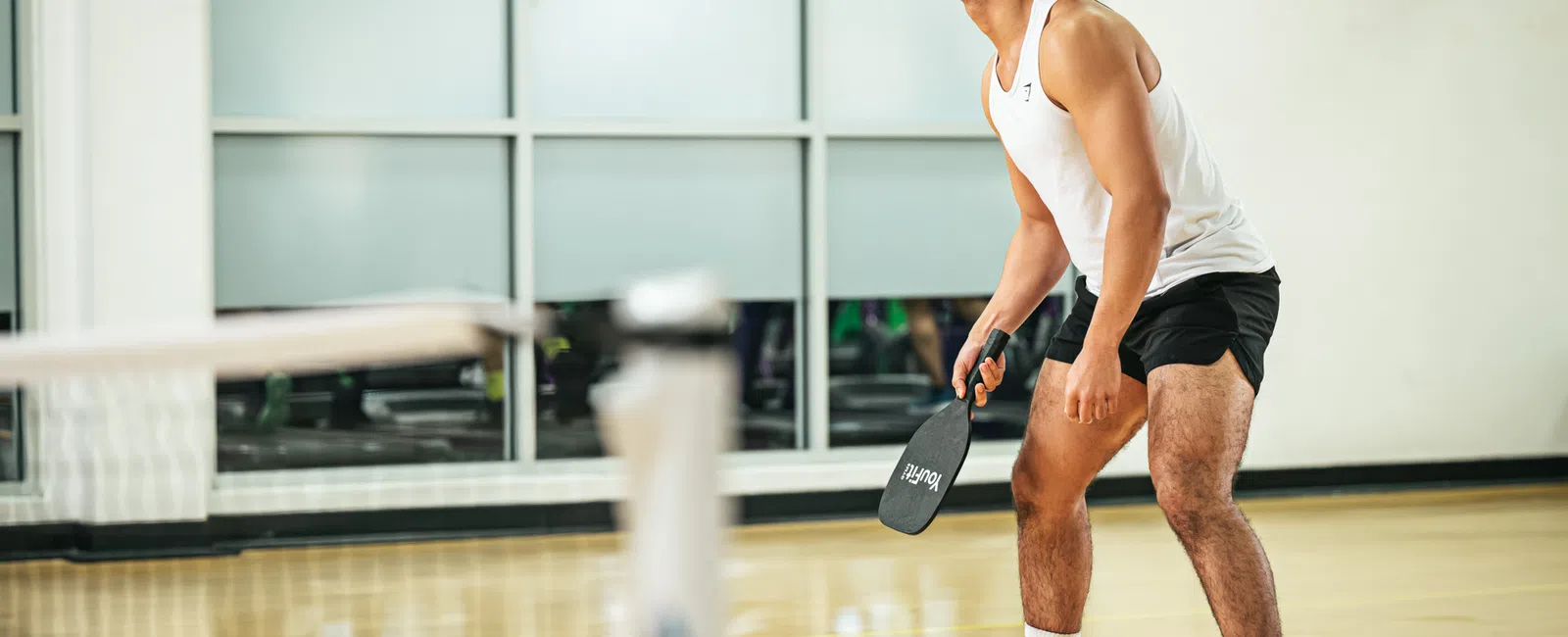 Athletic man in white tank top and black shorts playing pickleball indoors with paddle in hand.