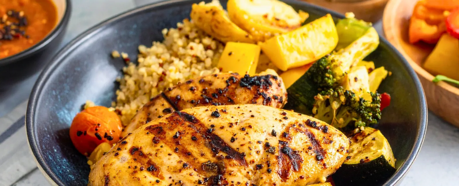 Grilled chicken breast with quinoa, roasted vegetables, and a sauce pitcher on a plate with side dishes.