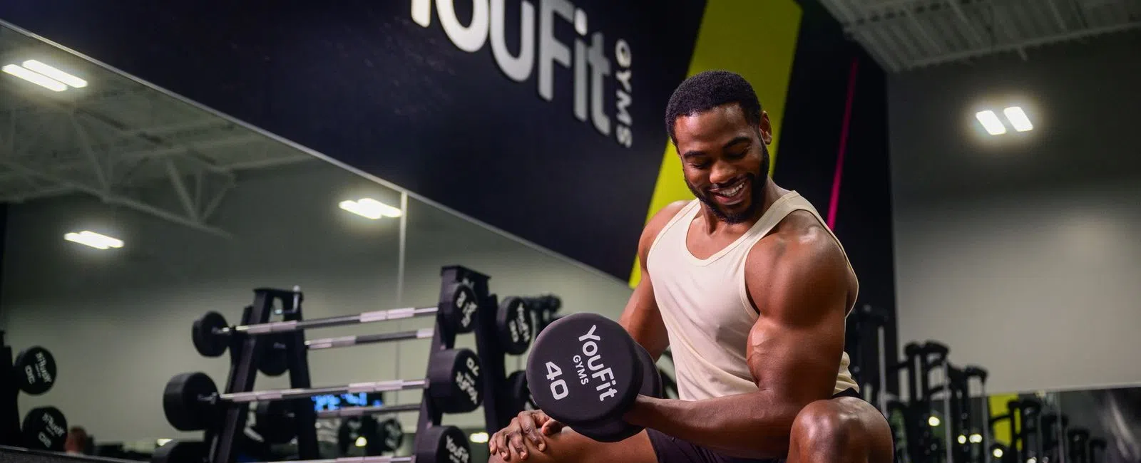 Smiling man lifting 40-pound dumbbell seated on bench in YouFit gym with weights and mirrored wall in background.