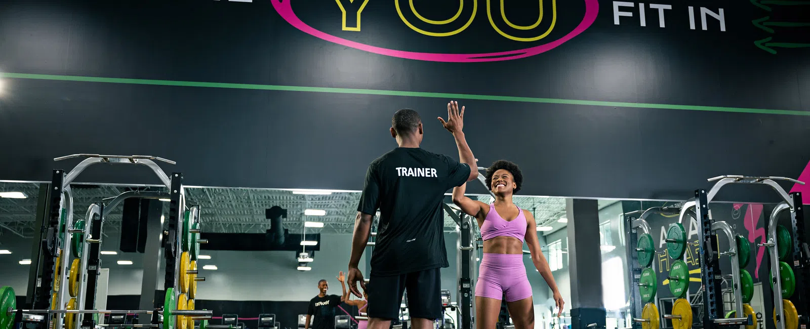Gym trainer and woman in purple workout clothes giving high five by weightlifting area under motivational sign.
