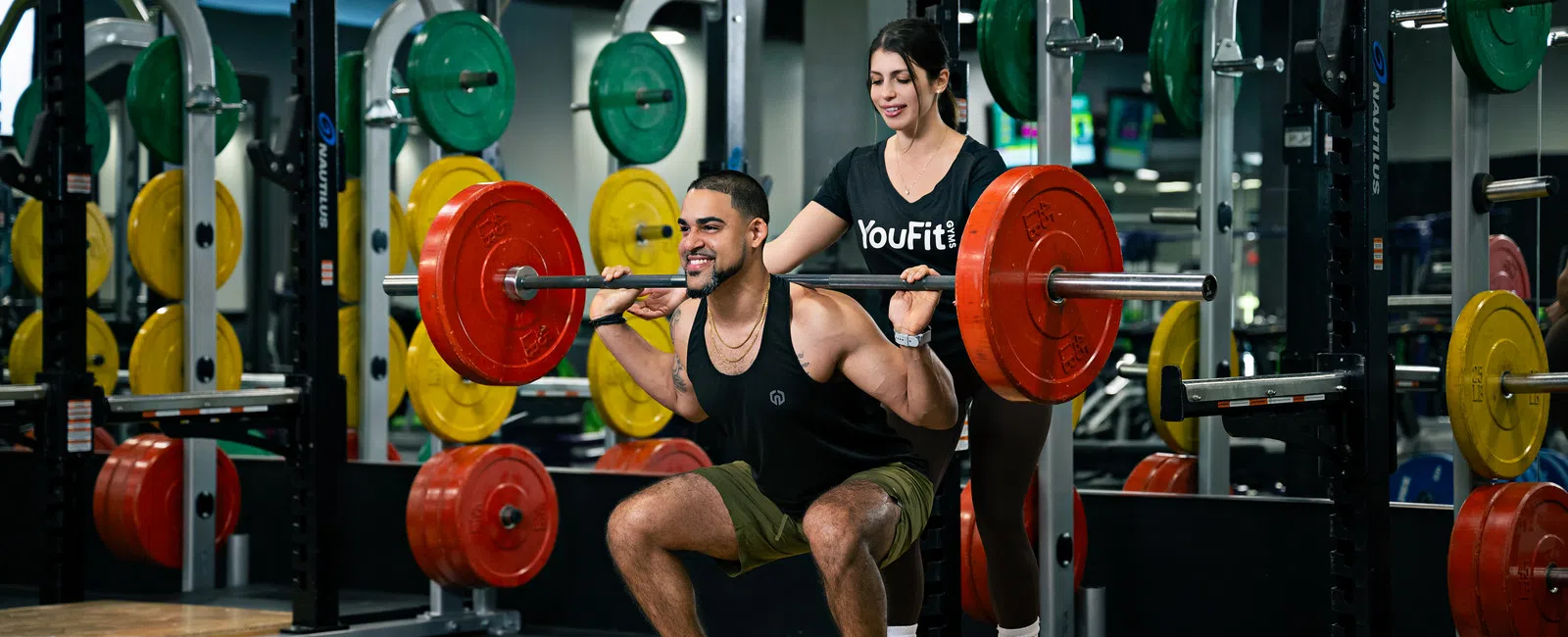 Man performs barbell squat with a trainer spotting him in a gym surrounded by colorful weight plates and mirrors.