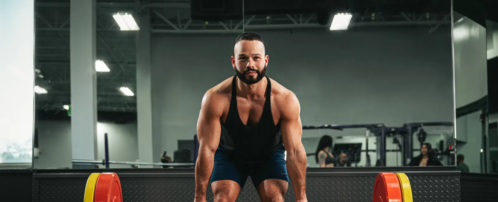 Muscular man wearing black tank top lifting heavy barbell with colorful weight plates in gym.